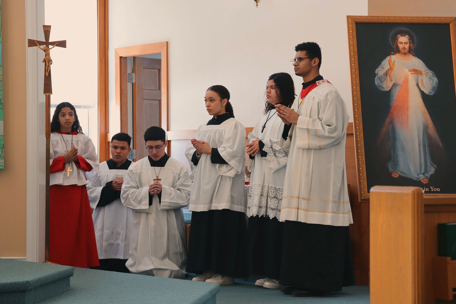 Altar servers wearing traditional white robes stand in prayer formation near a Divine Mercy image in a Catholic church.
