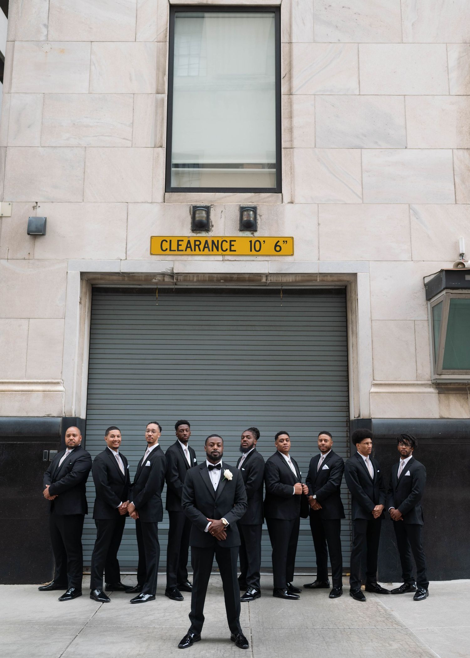 Wedding party members in black suits pose in front of an urban building with a clearance sign.
