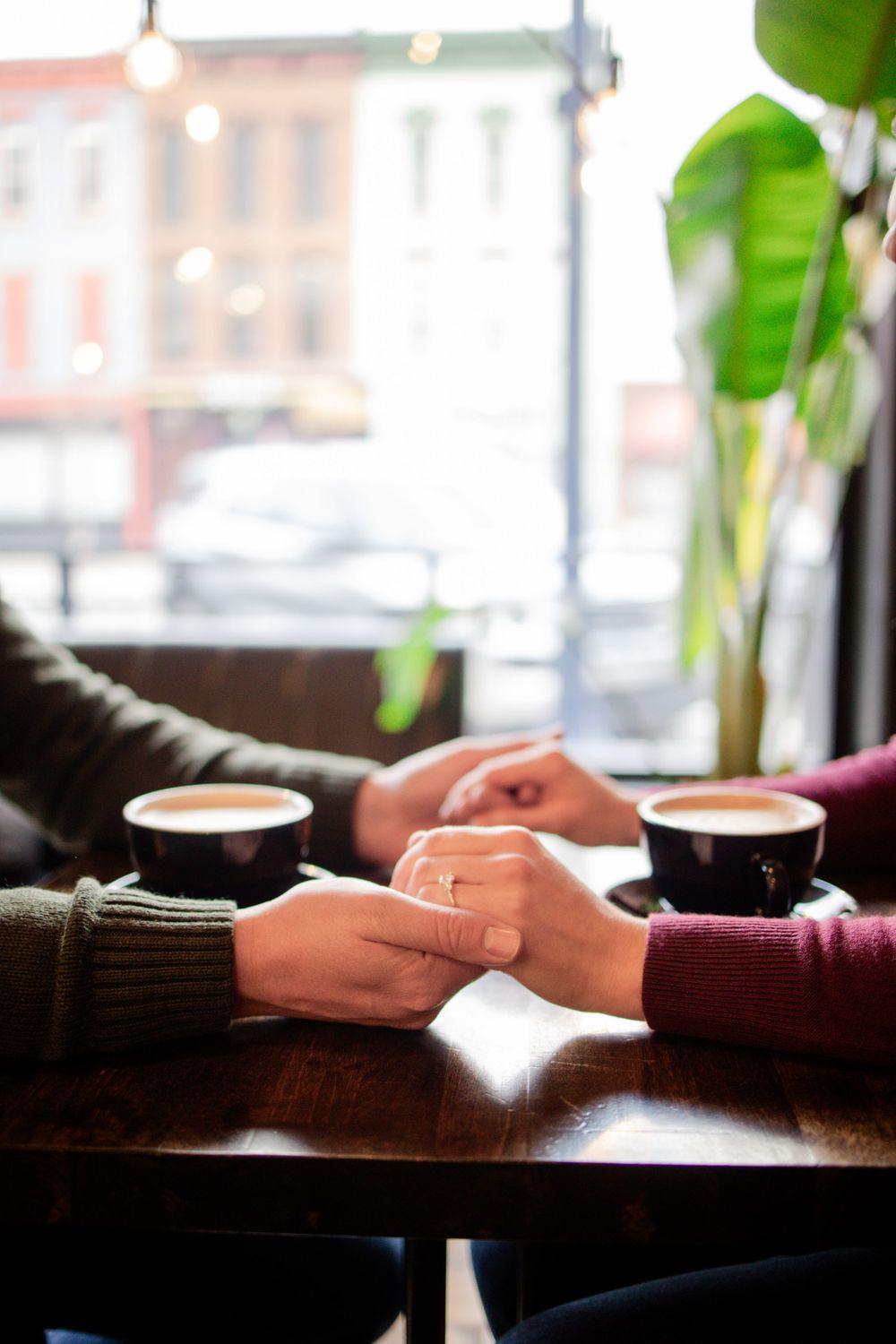 Two people holding hands across a cafe table with coffee cups and plants in background.