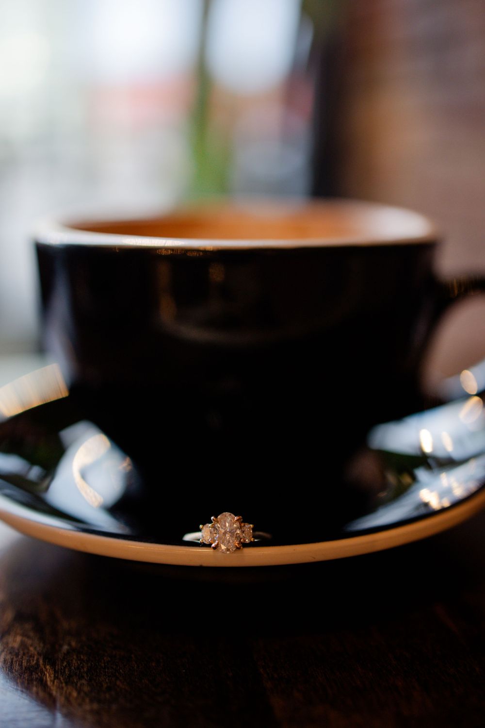 Black coffee cup on saucer with engagement ring displayed on rim.