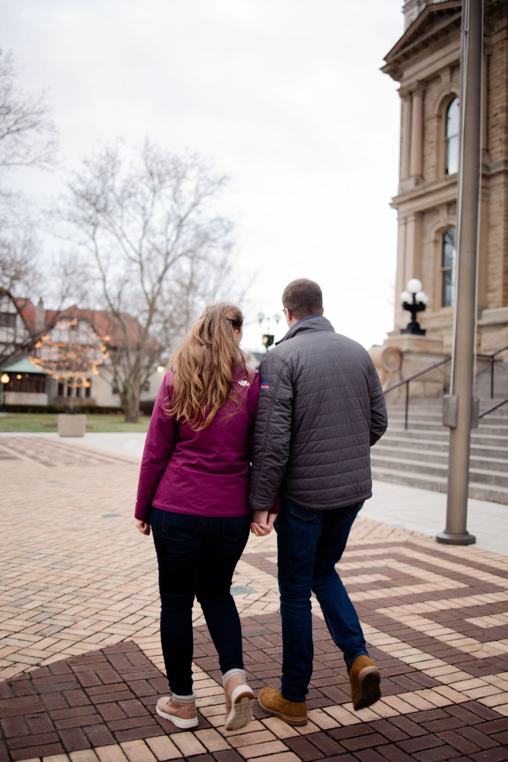 A couple walks together on a brick path towards a historic building in winter.
