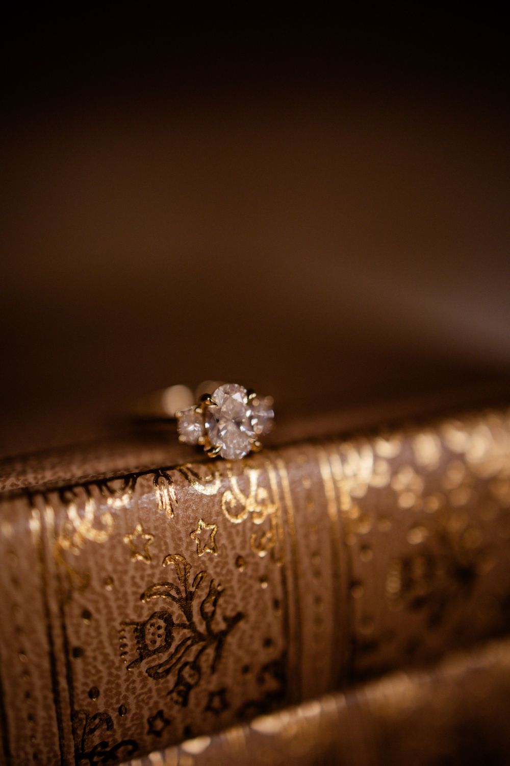 Diamond engagement ring sitting atop vintage ornate books in warm lighting.