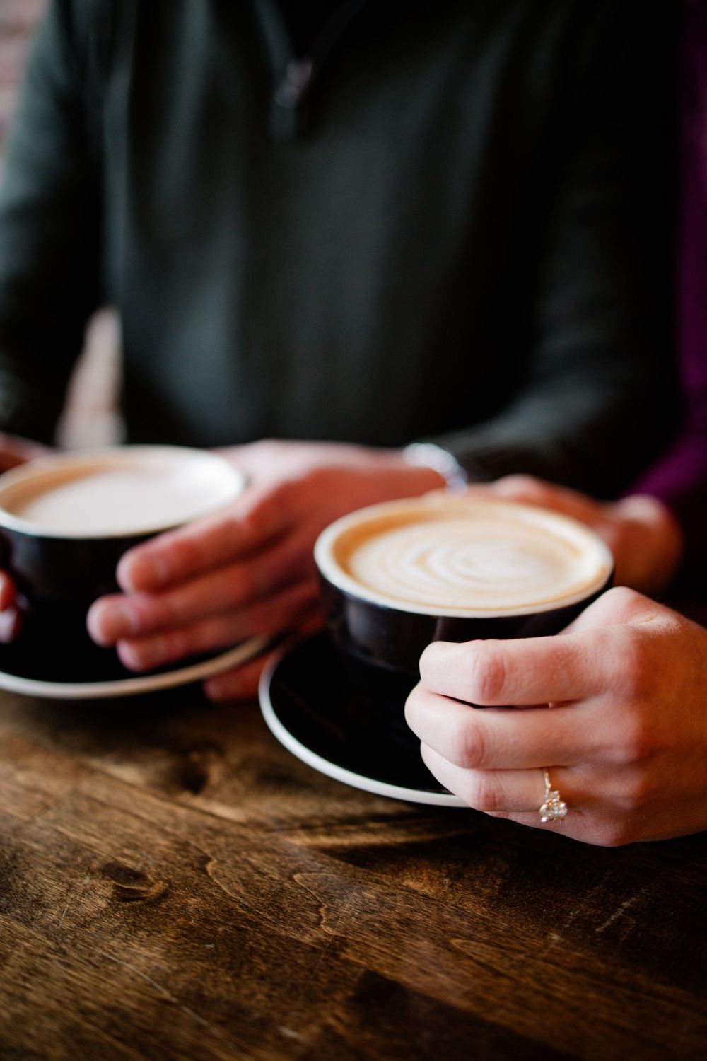 Hands holding two black coffee cups with latte art on a wooden table.