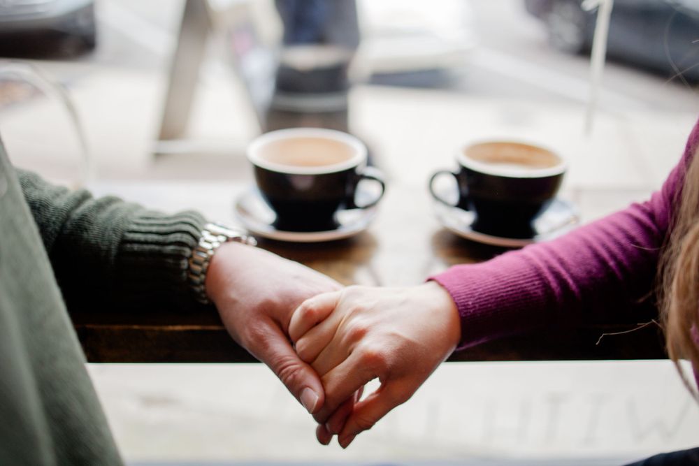 Two people holding hands across a coffee table with lattes in black cups.