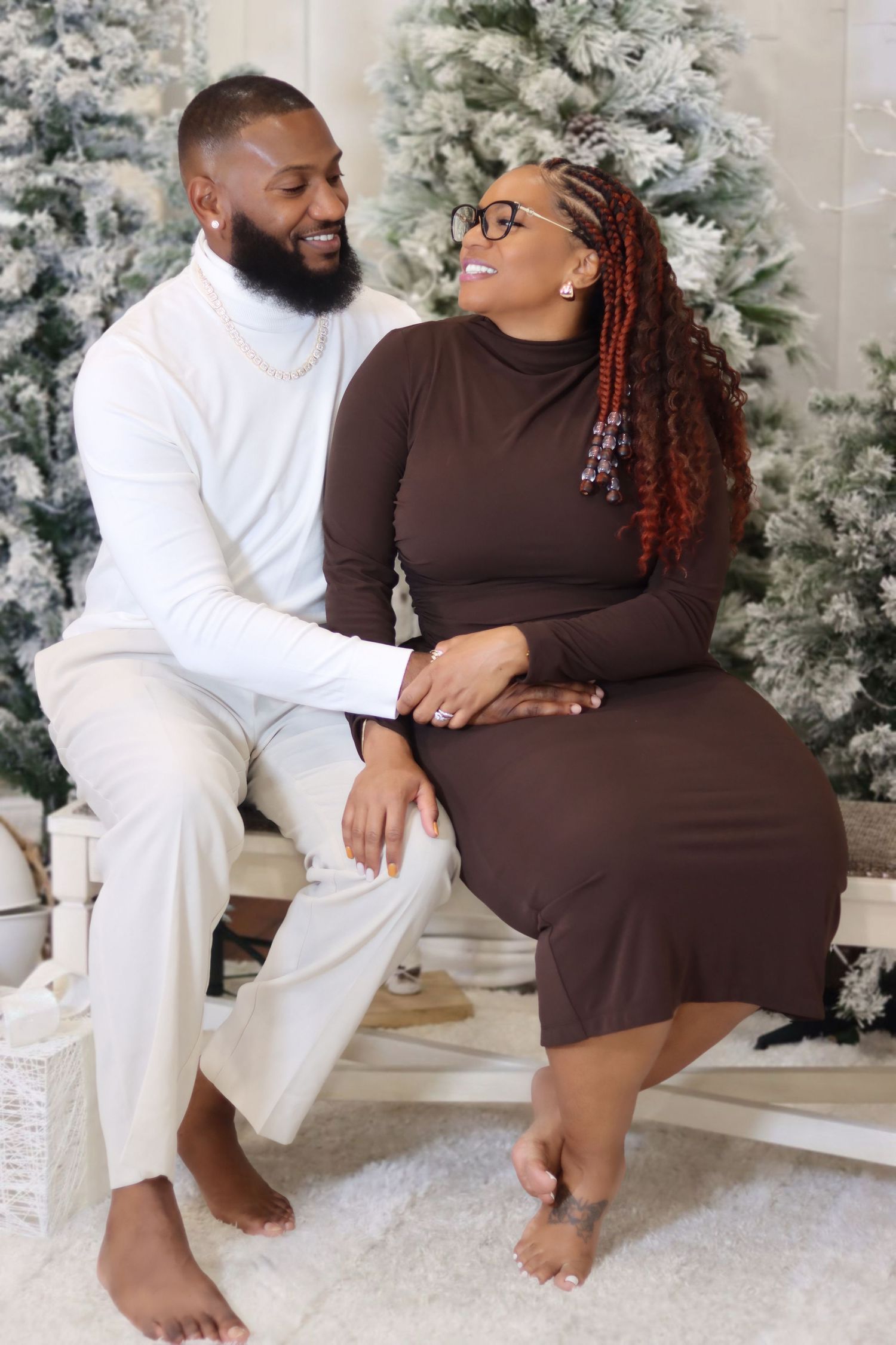 A couple shares a joyful moment together in front of snow-frosted Christmas trees wearing coordinating brown and white outfits.
