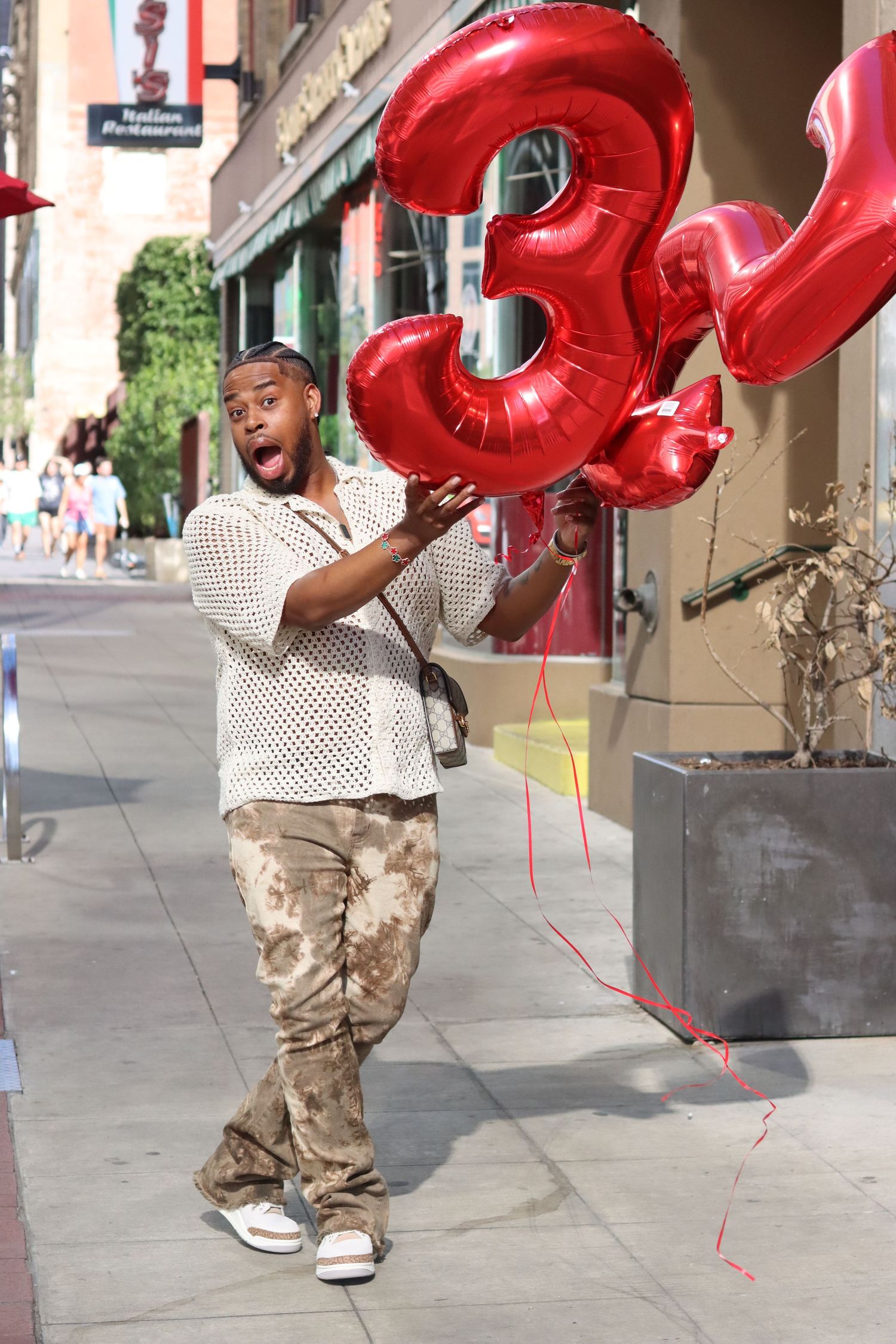 A person in camo pants and white shirt poses excitedly with large red number 3 balloons on a city street.