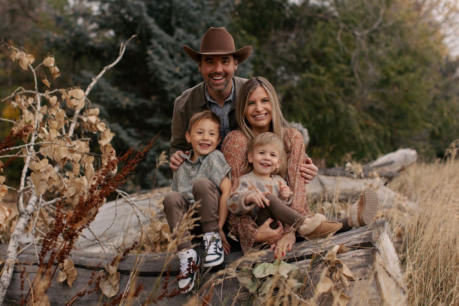 A family sits together on fallen logs in a natural setting with dried grasses and evergreen trees in the background.