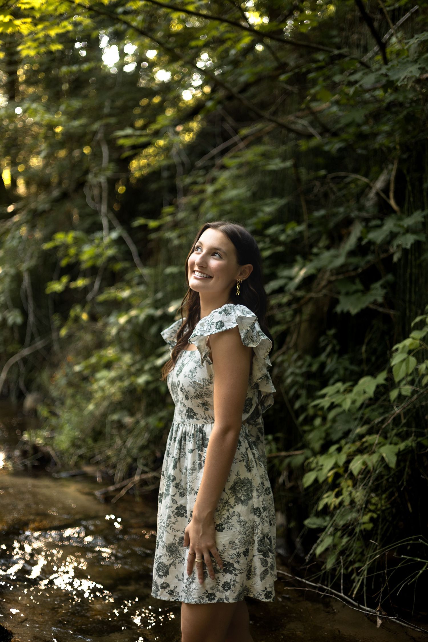 Woman in floral dress standing in a forest stream during golden hour with sunlight filtering through trees. West Michigan, Senior.