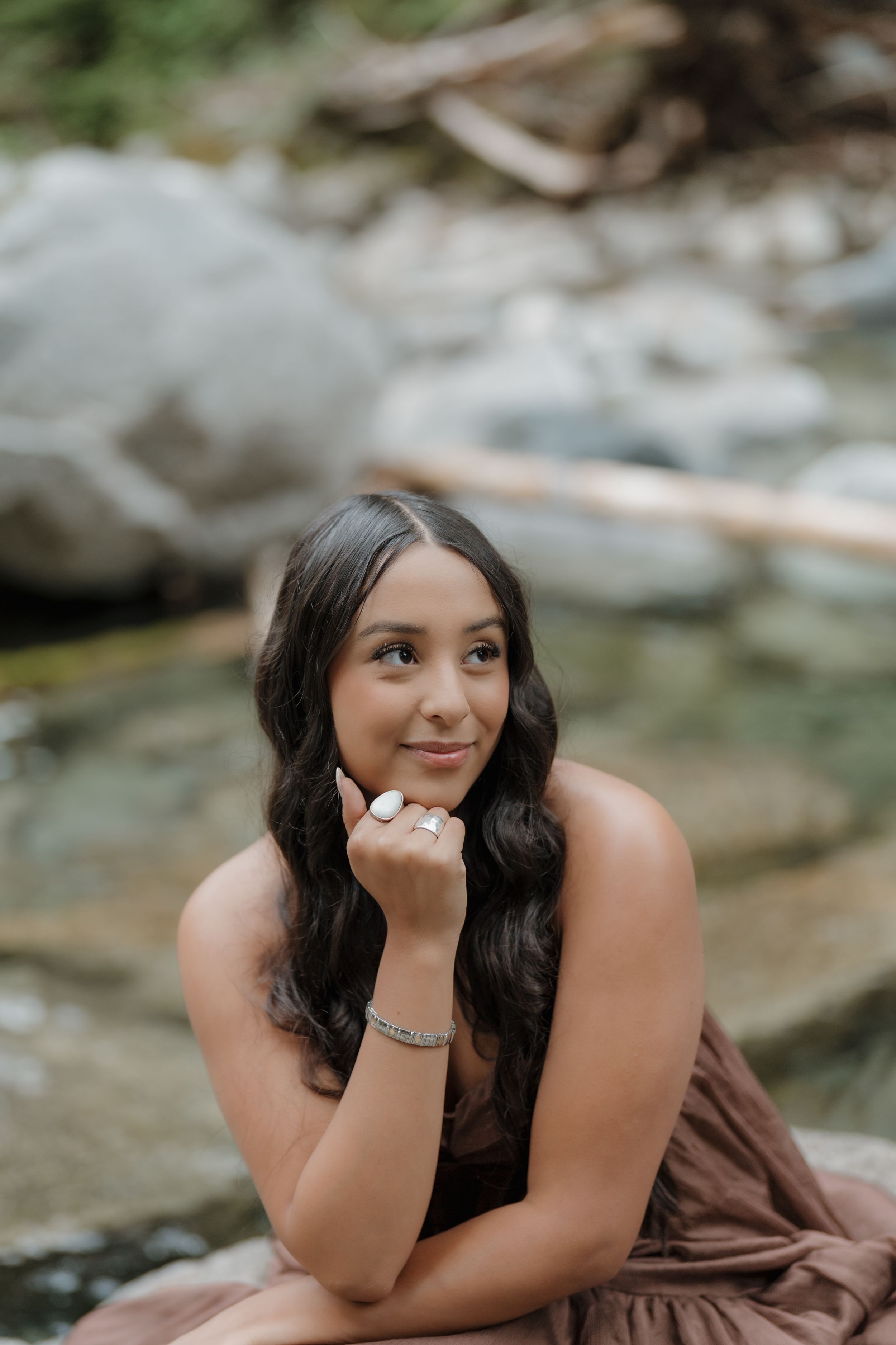 Young woman in brown dress poses gracefully on rocks by a stream, resting chin on hand, with long dark wavy hair.