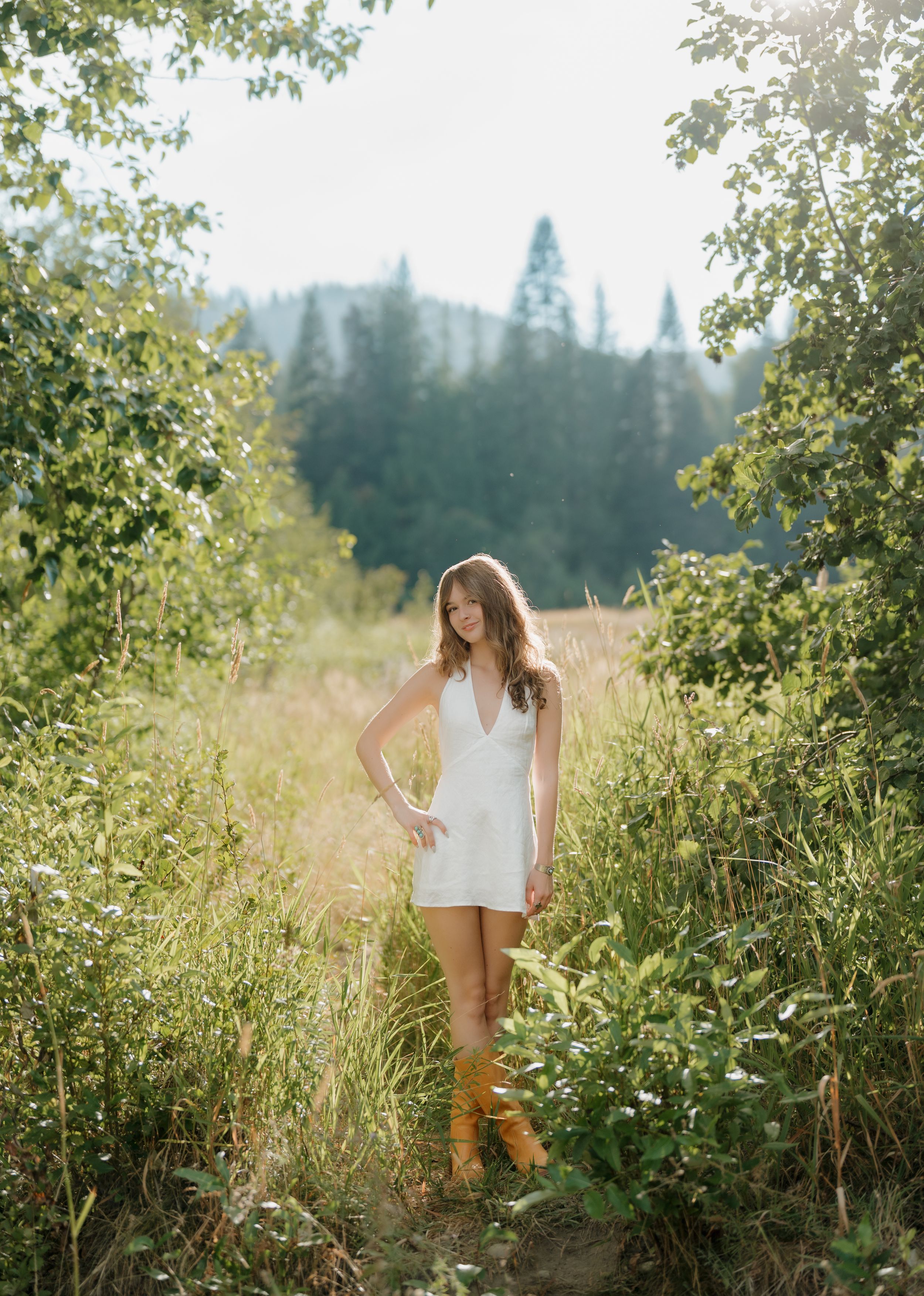 Young woman in white romper and tan boots poses in a sunlit forest meadow surrounded by lush green foliage.