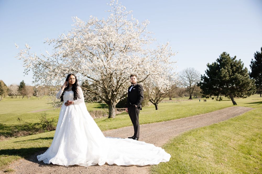 Bride in white gown and groom in suit stand on a path beside a blooming cherry blossom tree on a sunny day.