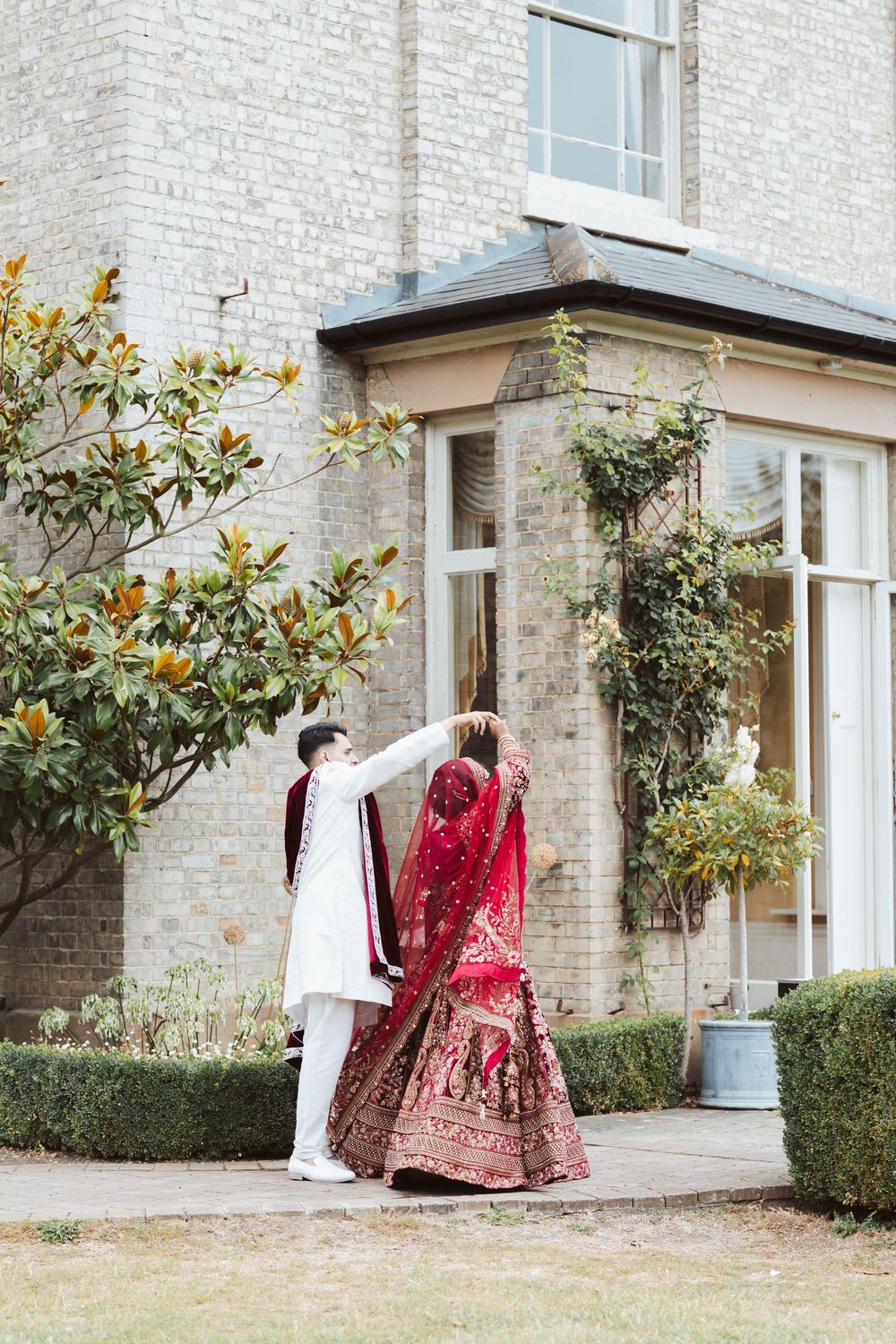 A traditional red and gold Indian bridal lehenga and white sherwani displayed outside a classic brick building.