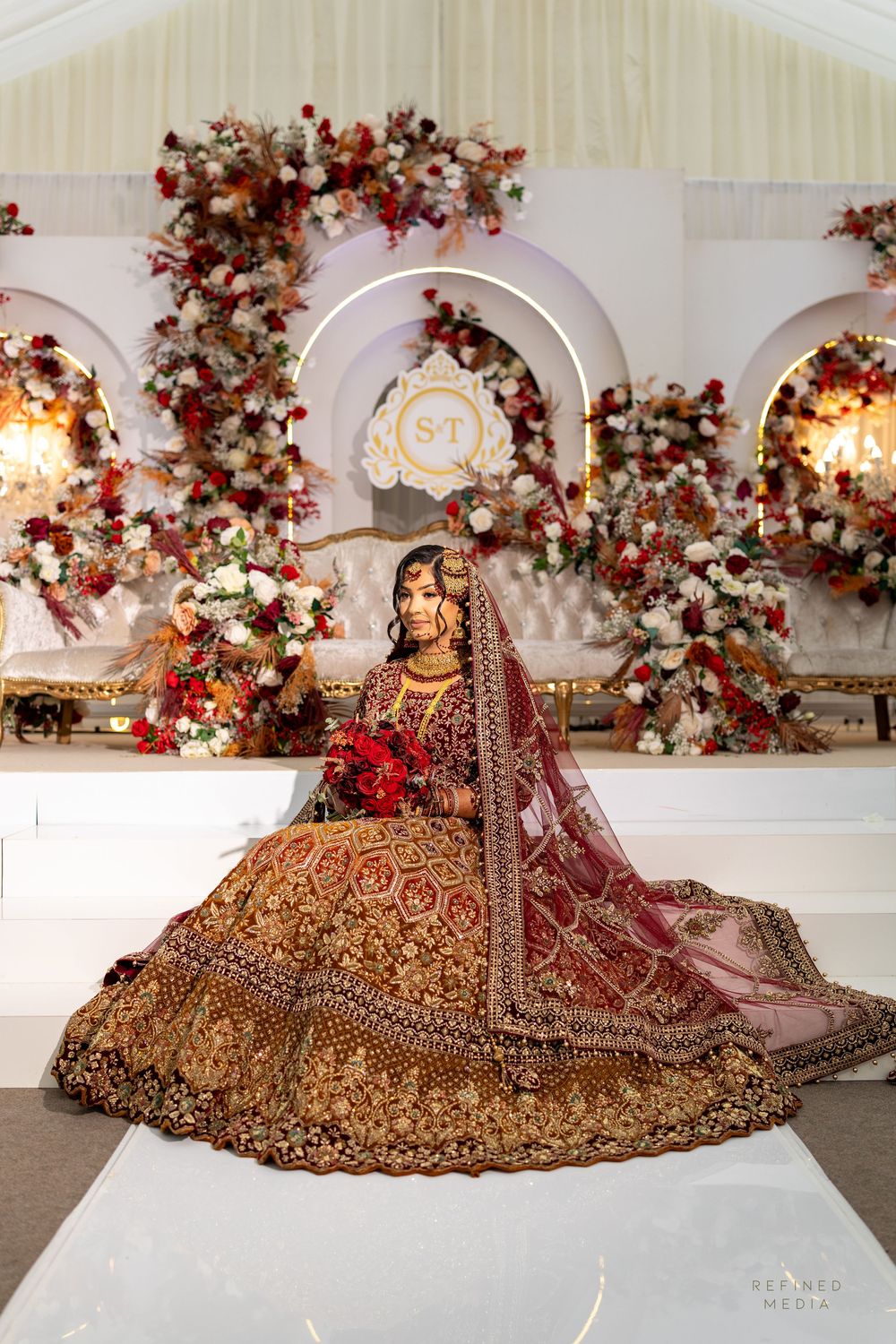 Luxurious burgundy and gold Indian bridal lehenga with intricate embroidery displayed in an elegant venue with floral decor.
