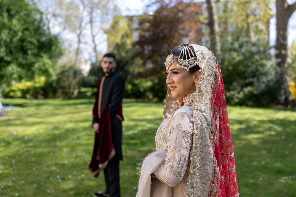 A bride in an ornate lace dress and red veil stands in a sunlit garden while a figure waits in the background.