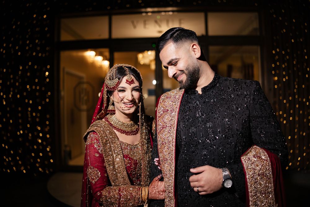 South Asian bride and groom in traditional wedding attire, smiling together outside a venue at night.