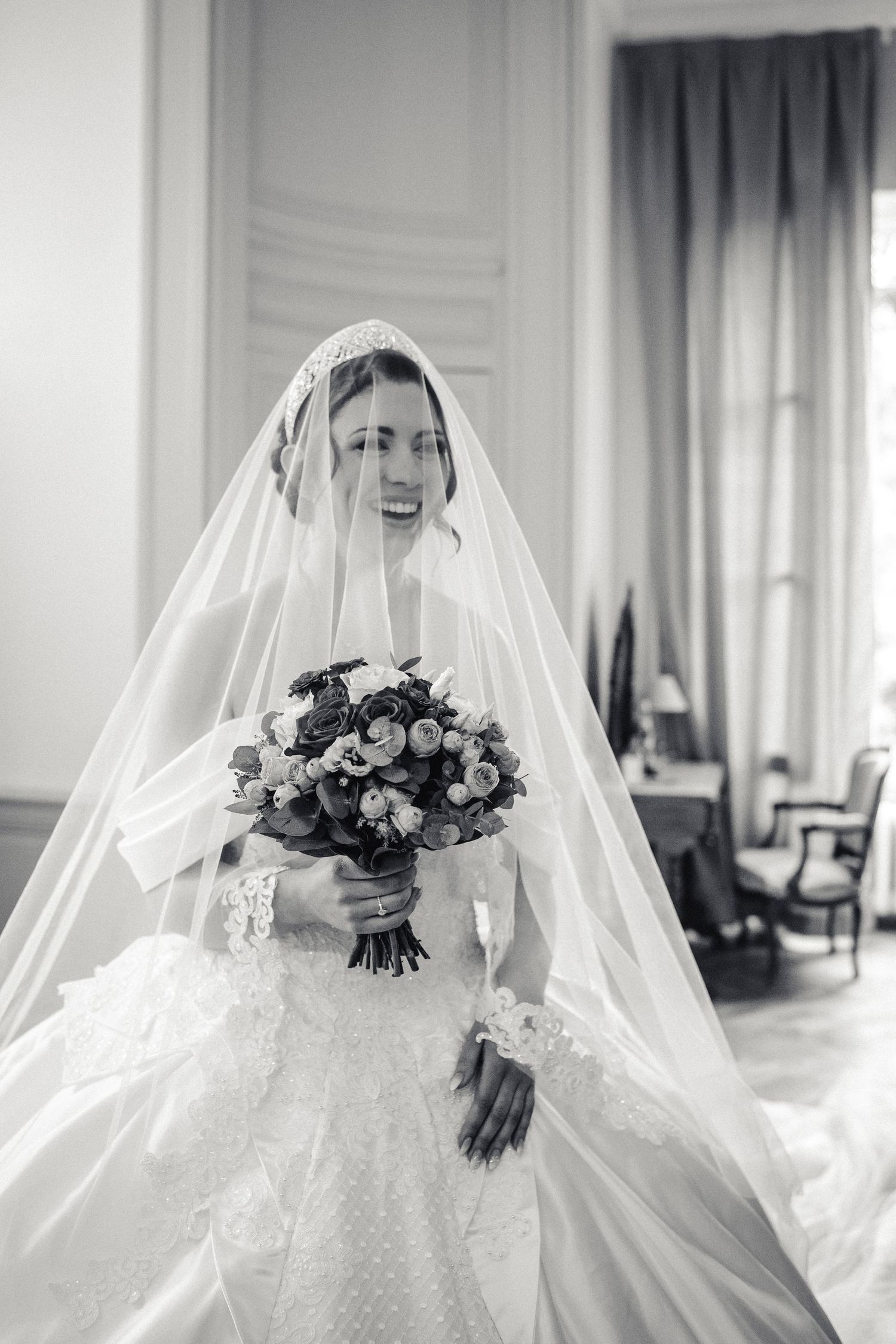 Mariée rayonnante en robe en dentelle et long voile, tenant un bouquet fleuri dans une pièce élégante d'un château.