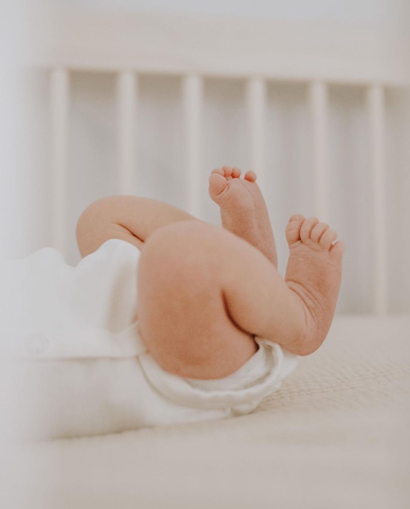 Close up of tiny baby feet and chubby legs resting on white bedding with crib railings in soft focus background.