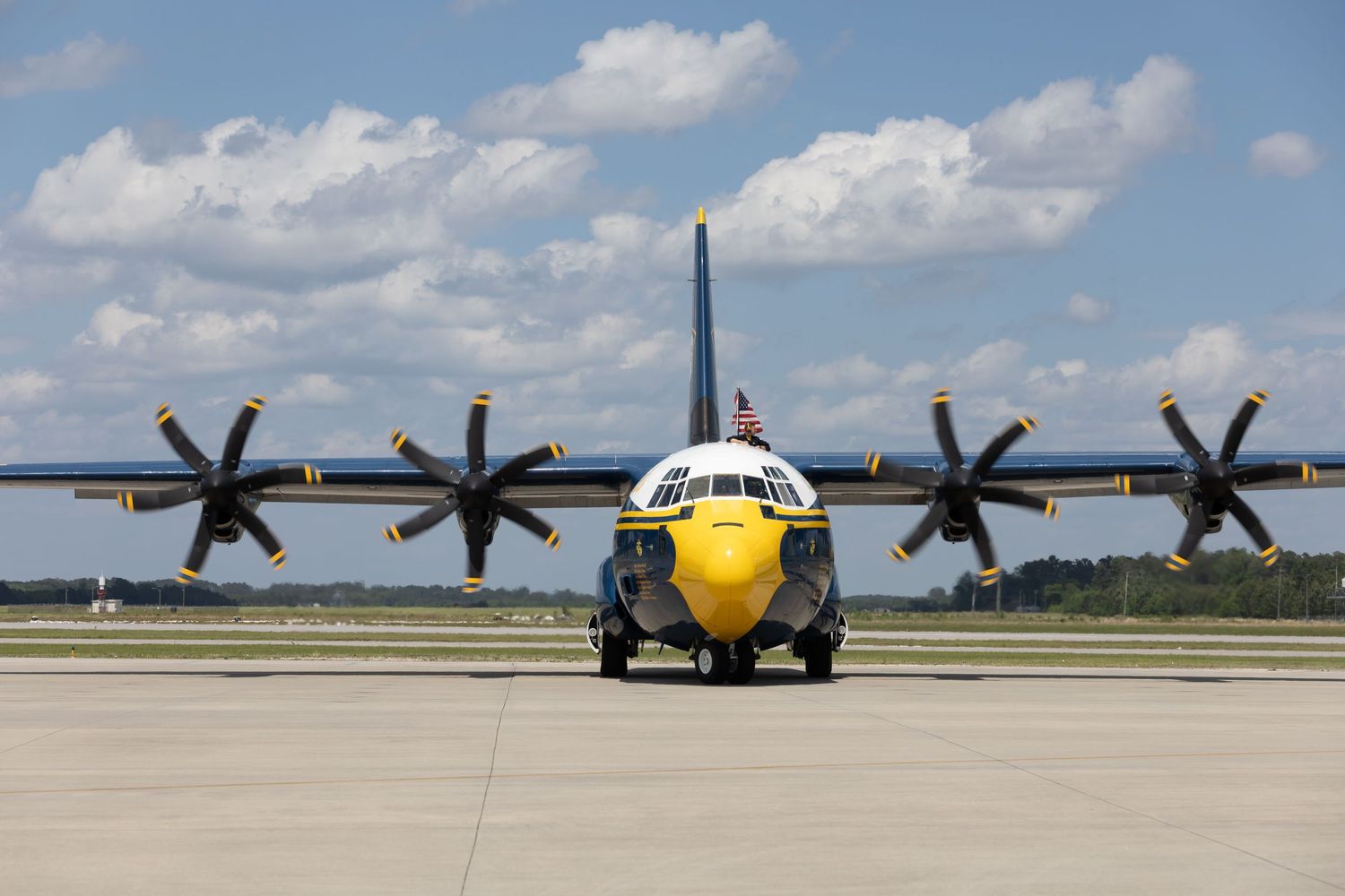A Blue Angels C-130 transport aircraft with yellow nose cone sits on tarmac under puffy white clouds.