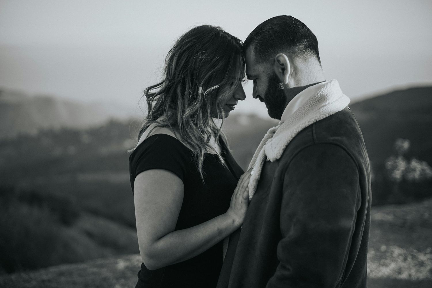A romantic black and white photo shows a couple sharing an intimate moment on a hilltop with a misty landscape behind them.
