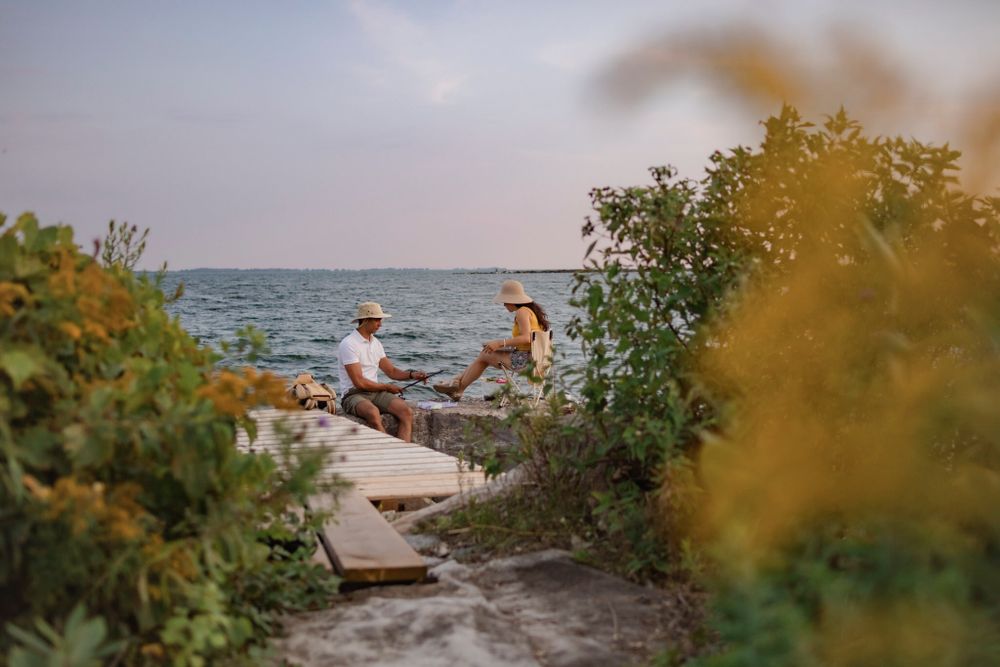 Couple sits on a breakwall of Lake Ontario preparing to go fishing. He baits his hook while she sits in a lawn chair and reads. Kingston, ON