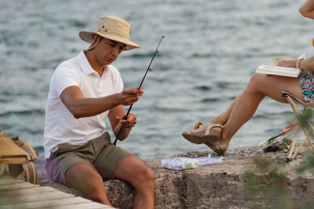 Couple sits on a breakwall of Lake Ontario preparing to go fishing. He baits his hook while she sits in a lawn chair and reads. Kingston, ON