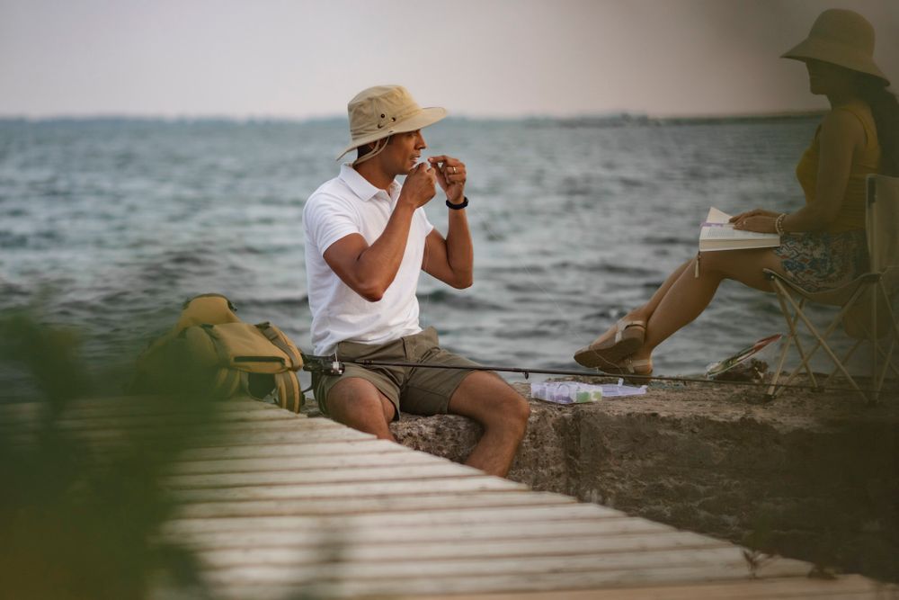 Couple sits on a breakwall of Lake Ontario preparing to go fishing. He baits his hook while she sits in a lawn chair and reads. Kingston, ON