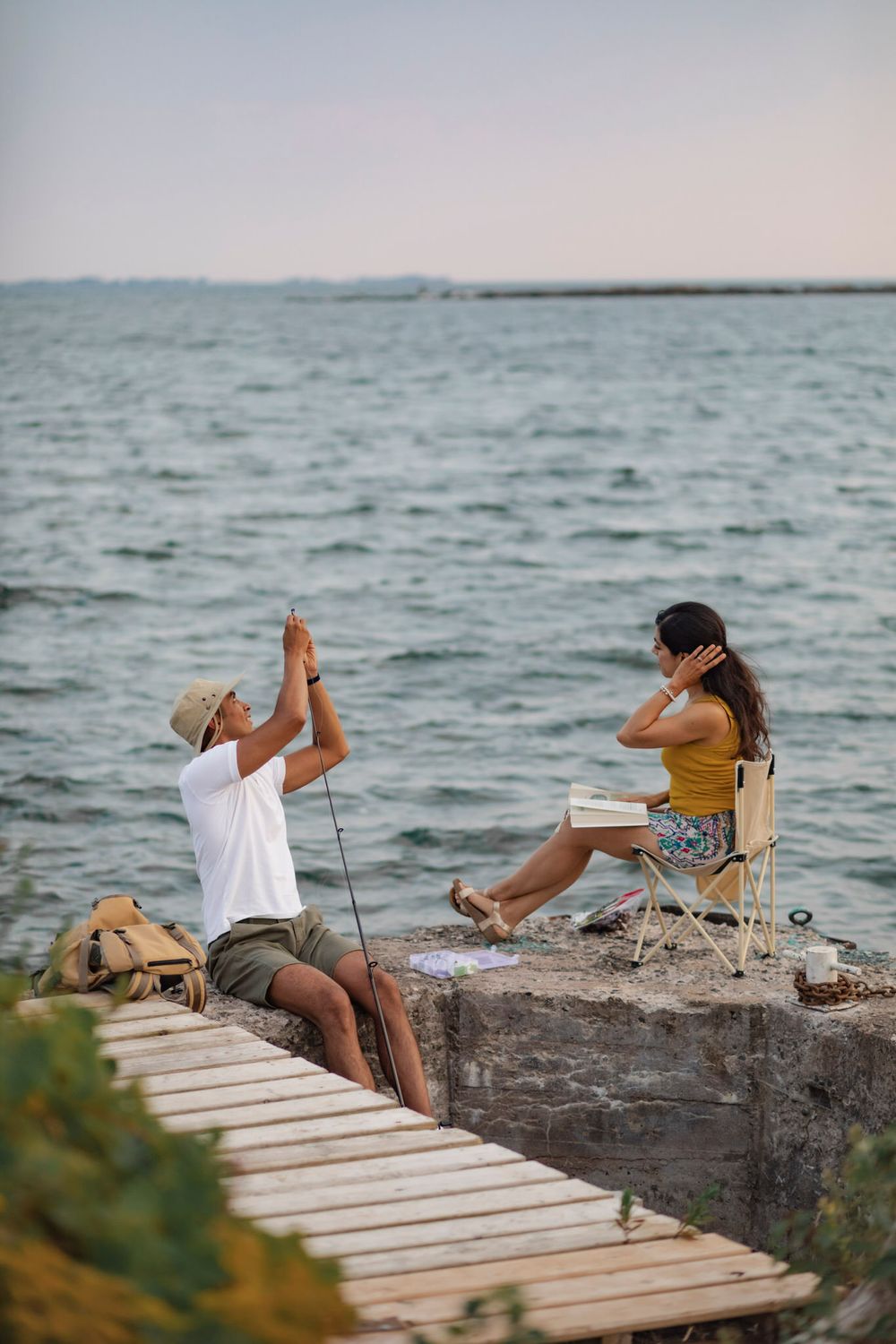 Couple sits on a breakwall of Lake Ontario preparing to go fishing. He baits his hook while she sits in a lawn chair and reads. Kingston, ON