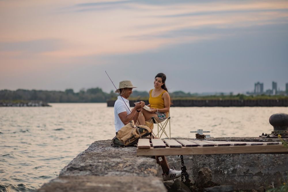 Couple sits on a breakwall of Lake Ontario preparing to go fishing. He baits his hook while she sits in a lawn chair and reads. Kingston, ON