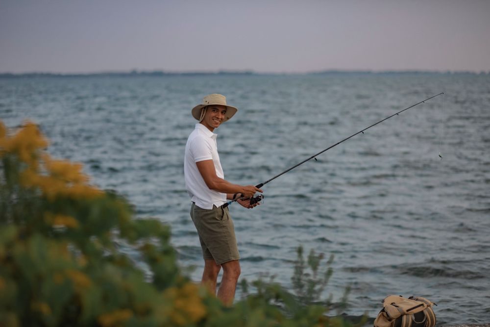 Man fishes from the shoreline of Lake Ontario. He looks over his shoulder and past greenery and smiles.