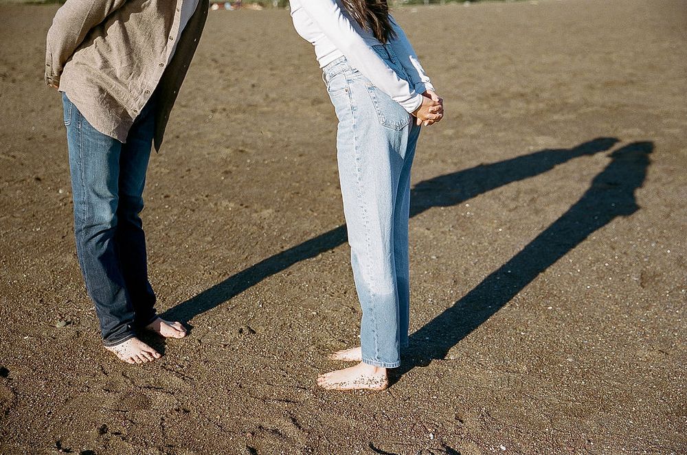 Two people standing on dirt ground casting long shadows while wearing jeans and casual footwear.