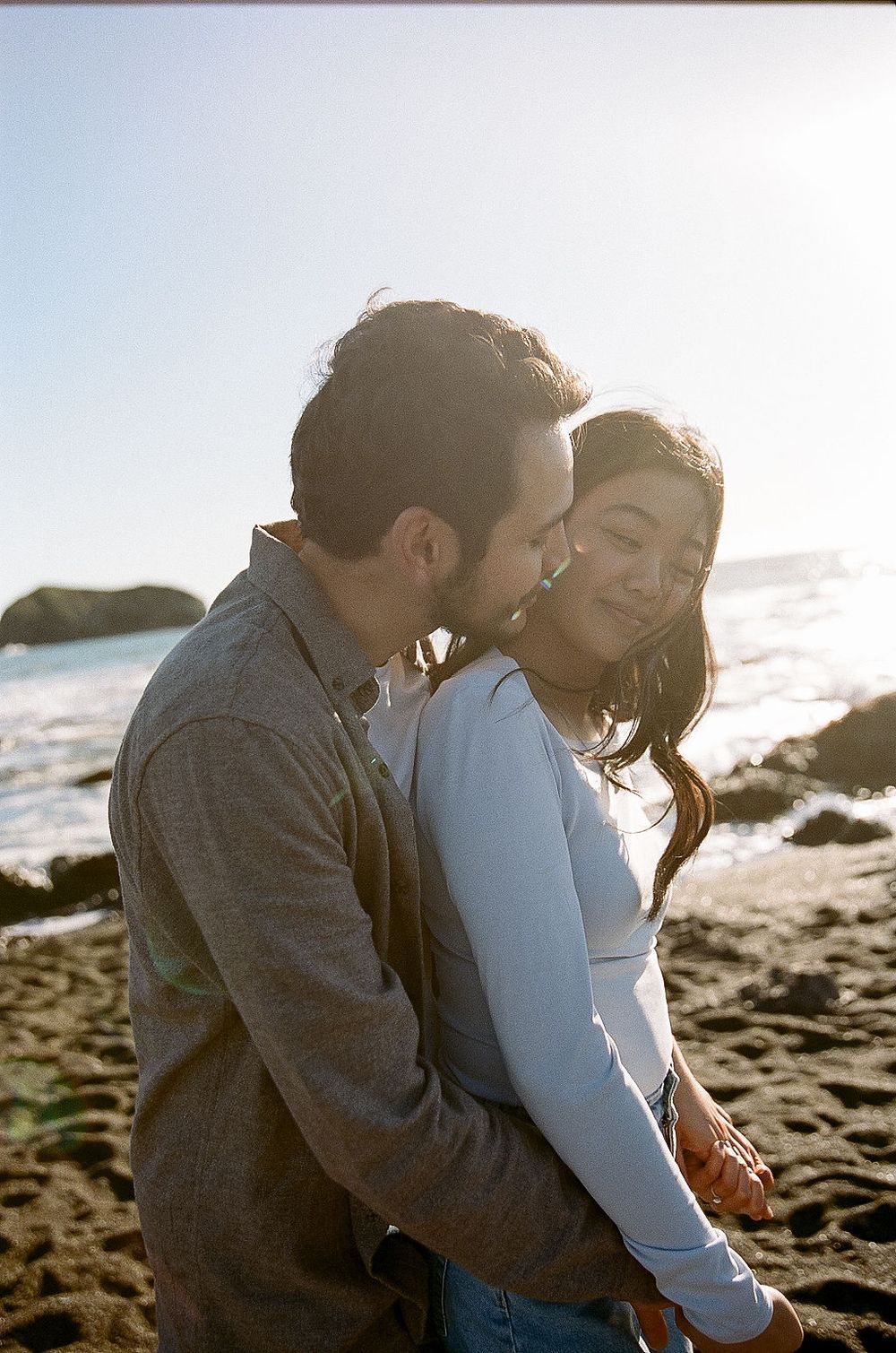 A romantic couple shares an intimate moment on a beach at sunset with ocean waves and rocks in the background.