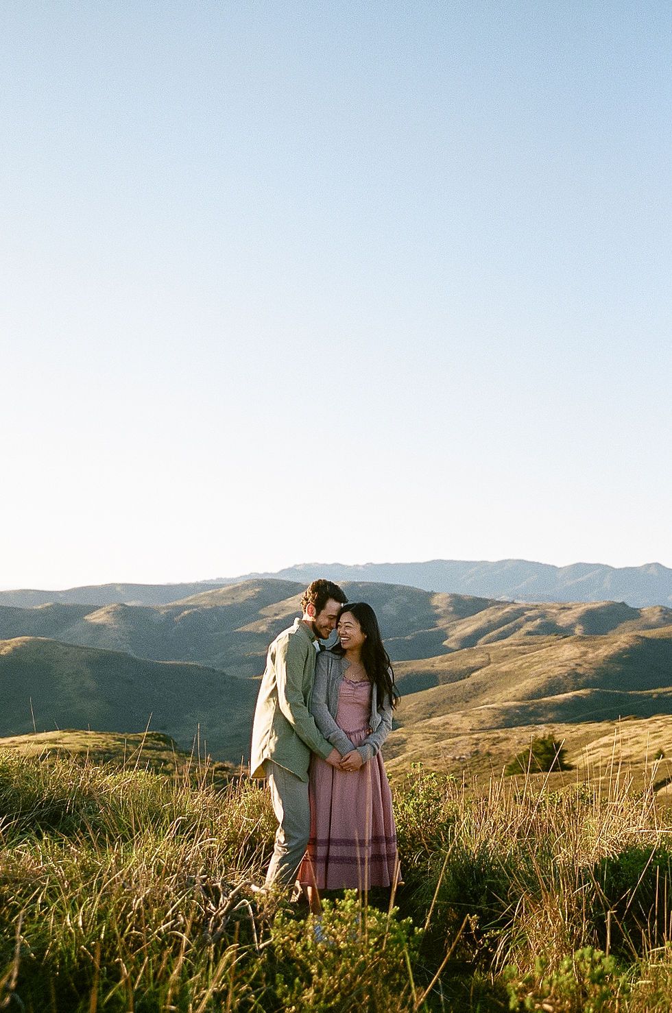 A couple embraces in golden sunlight on a hillside with rolling mountains stretching into the distance.