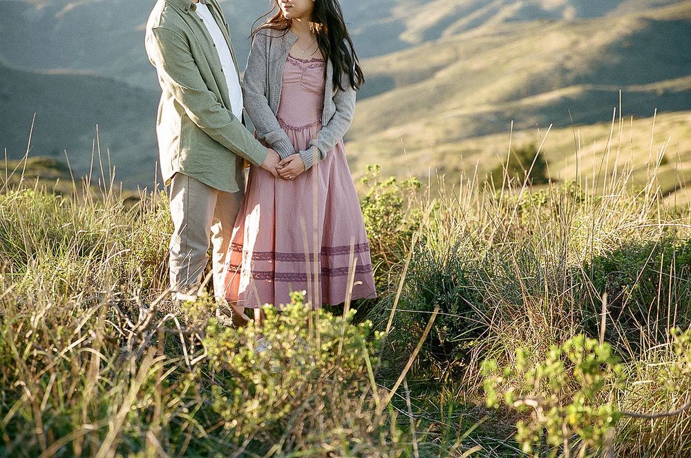 A couple holding hands in a scenic meadow with rolling hills and golden light in the background.