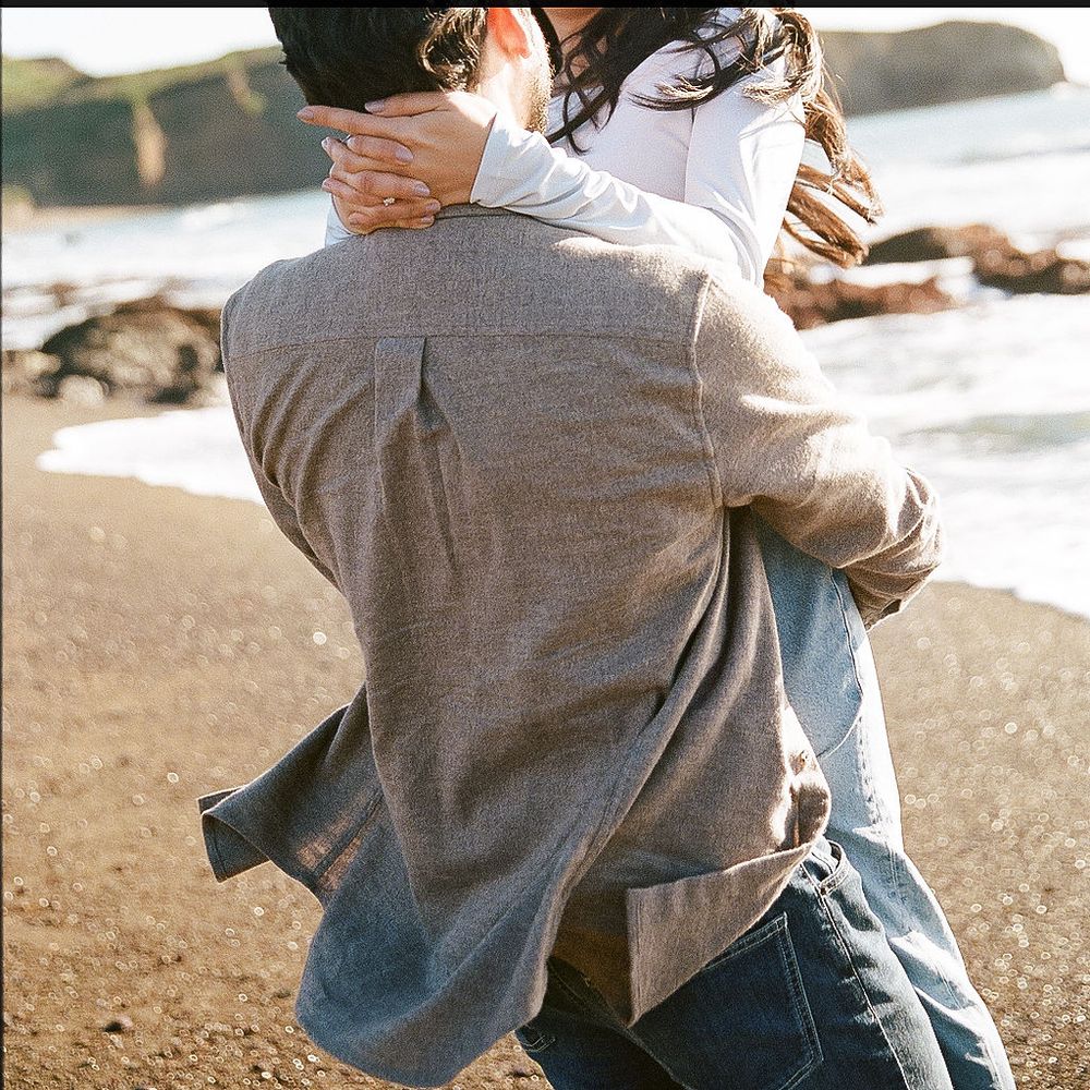Someone wearing a gray jacket and blue jeans embracing another person on a sunny beach.
