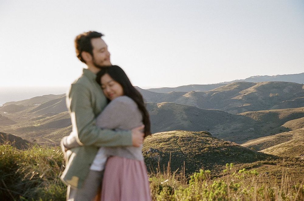 A couple embraces while enjoying a scenic view of rolling hills and mountains during golden hour.