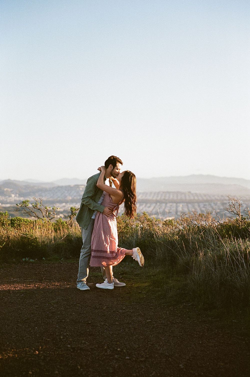 A romantic couple embraces on a hilltop trail at sunset with rolling hills and mountains in the background.