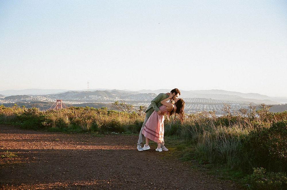 A couple embraces on a hilltop trail overlooking a scenic bay landscape during golden hour.