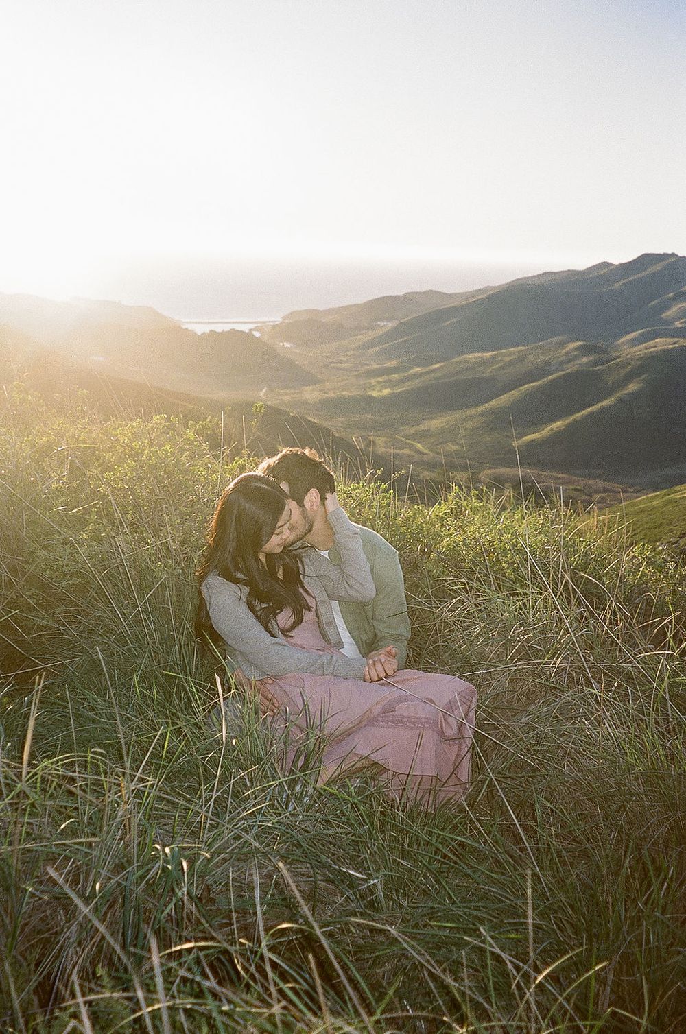 An intimate moment between a couple sitting in tall grass during sunset with mountain views.
