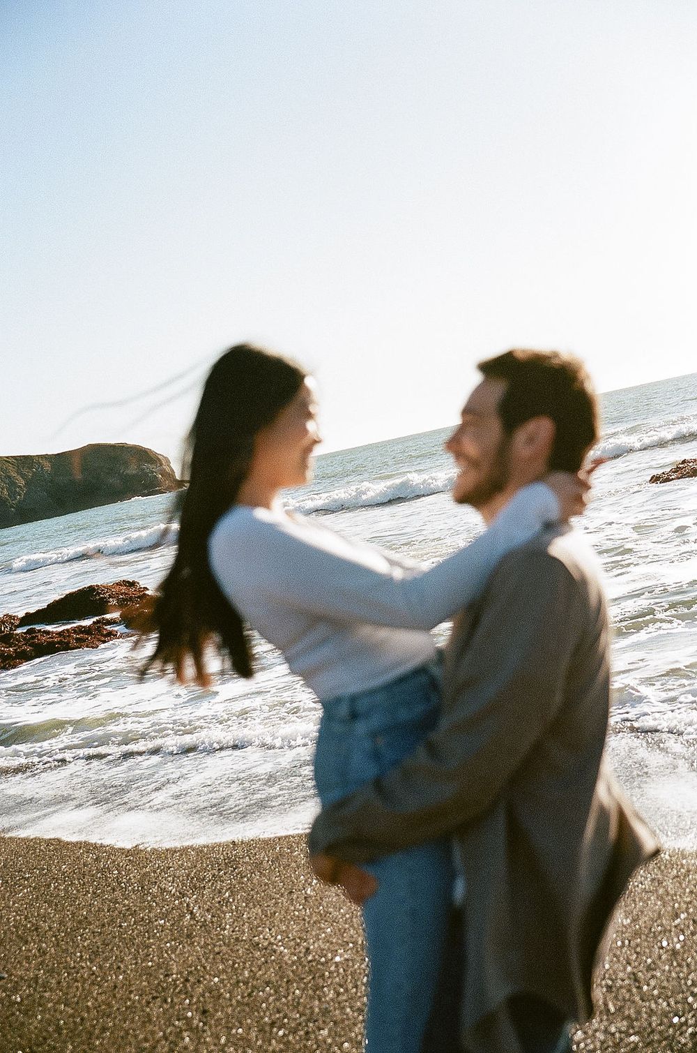 A couple embraces on a sunny beach with waves crashing against rocky shores in the background.