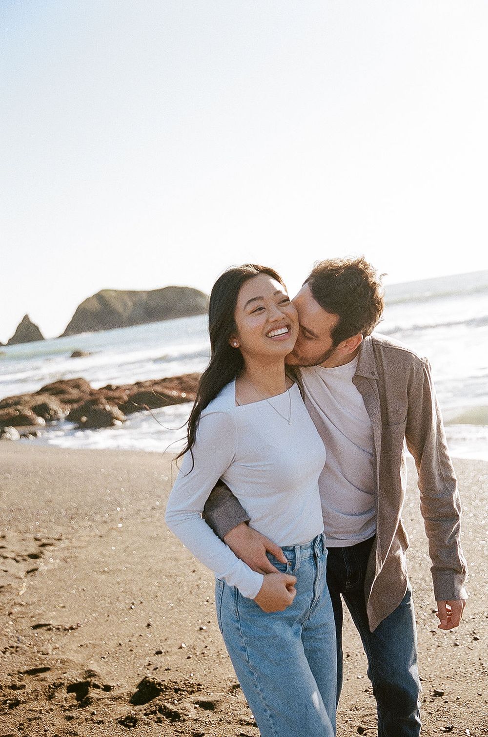 A romantic couple shares a joyful moment together on a sunny beach with coastal rock formations in the background.