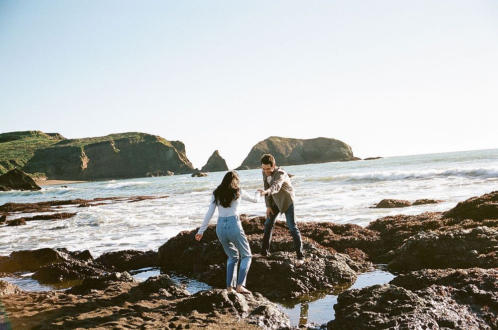 Two people explore rocky coastal cliffs along the ocean shore on a sunny day as waves crash against the rocks.