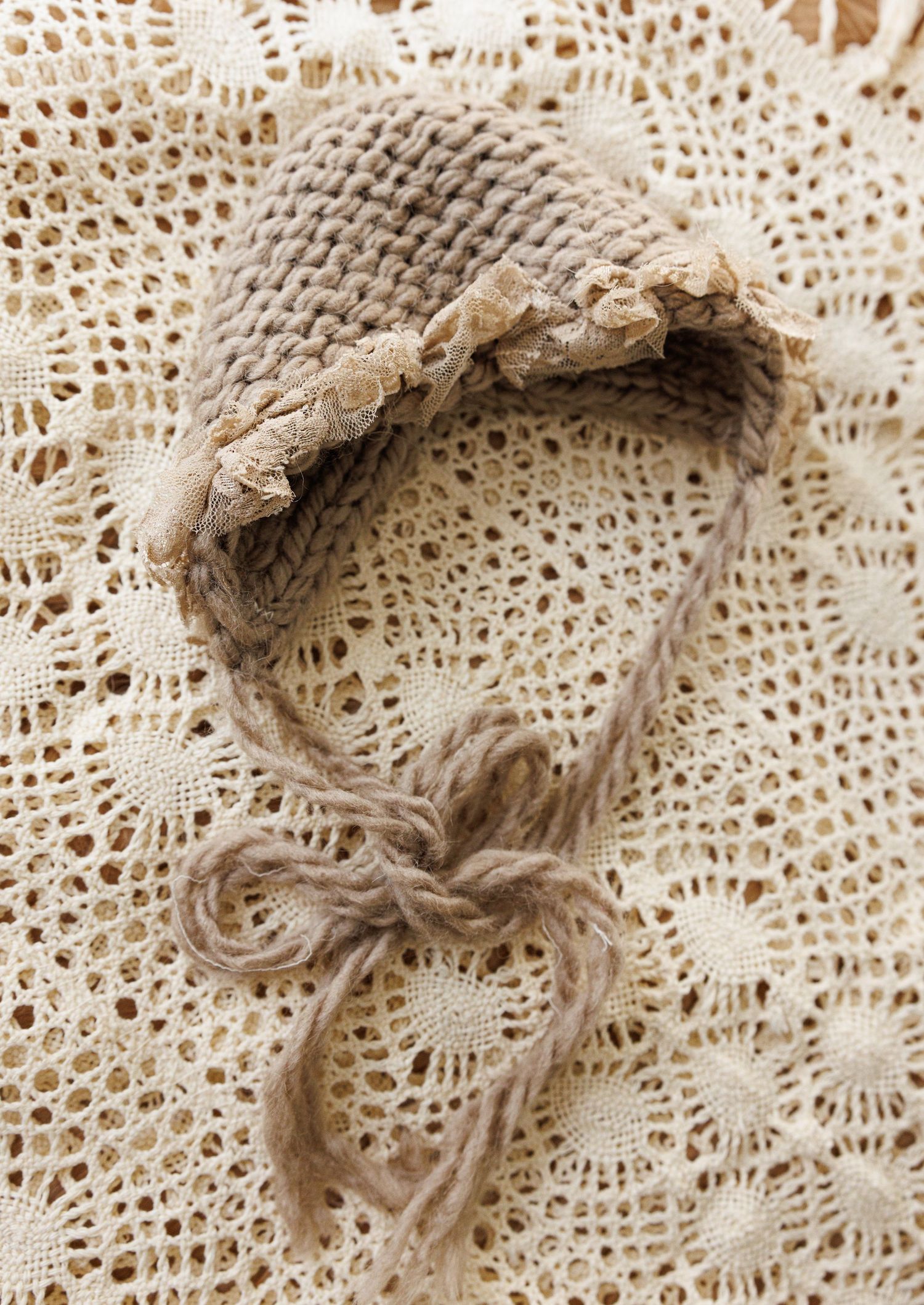 Vintage knitted baby bonnet with lace trim and ties rests on delicate crocheted doily background in neutral beige tones.