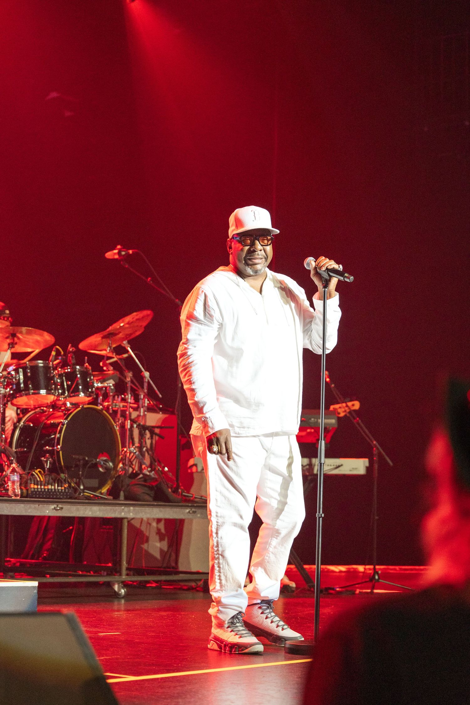 Performer in an all-white outfit singing on stage with red lighting and musical equipment in the background.
