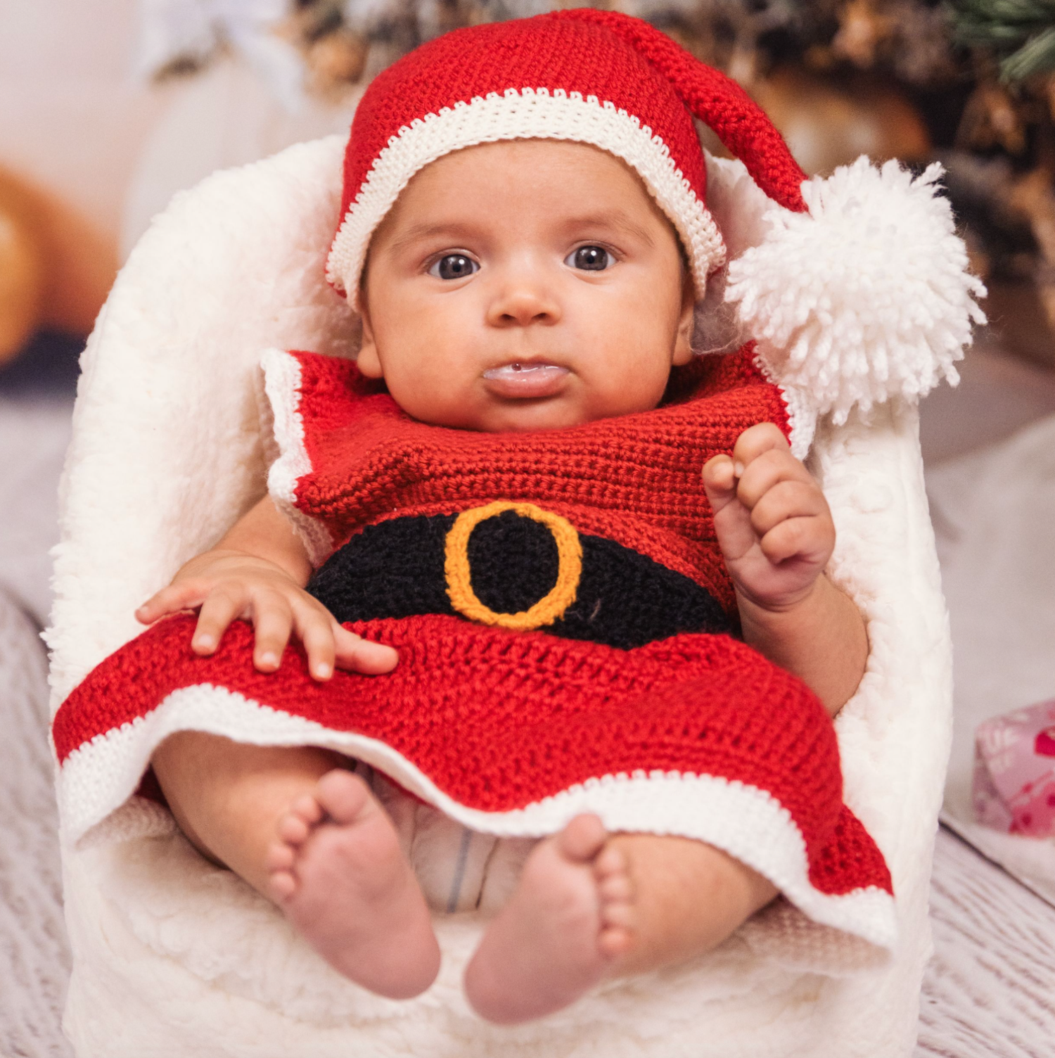 Baby wearing red and white crocheted Santa outfit lying on white blanket.