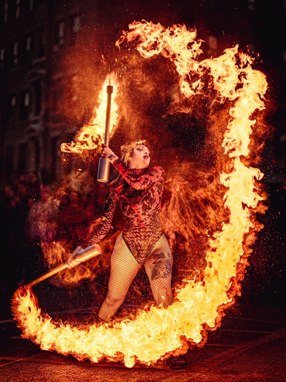 Fire performer in red costume twirling flaming batons creating a large circular fire arc against a dark night sky.
