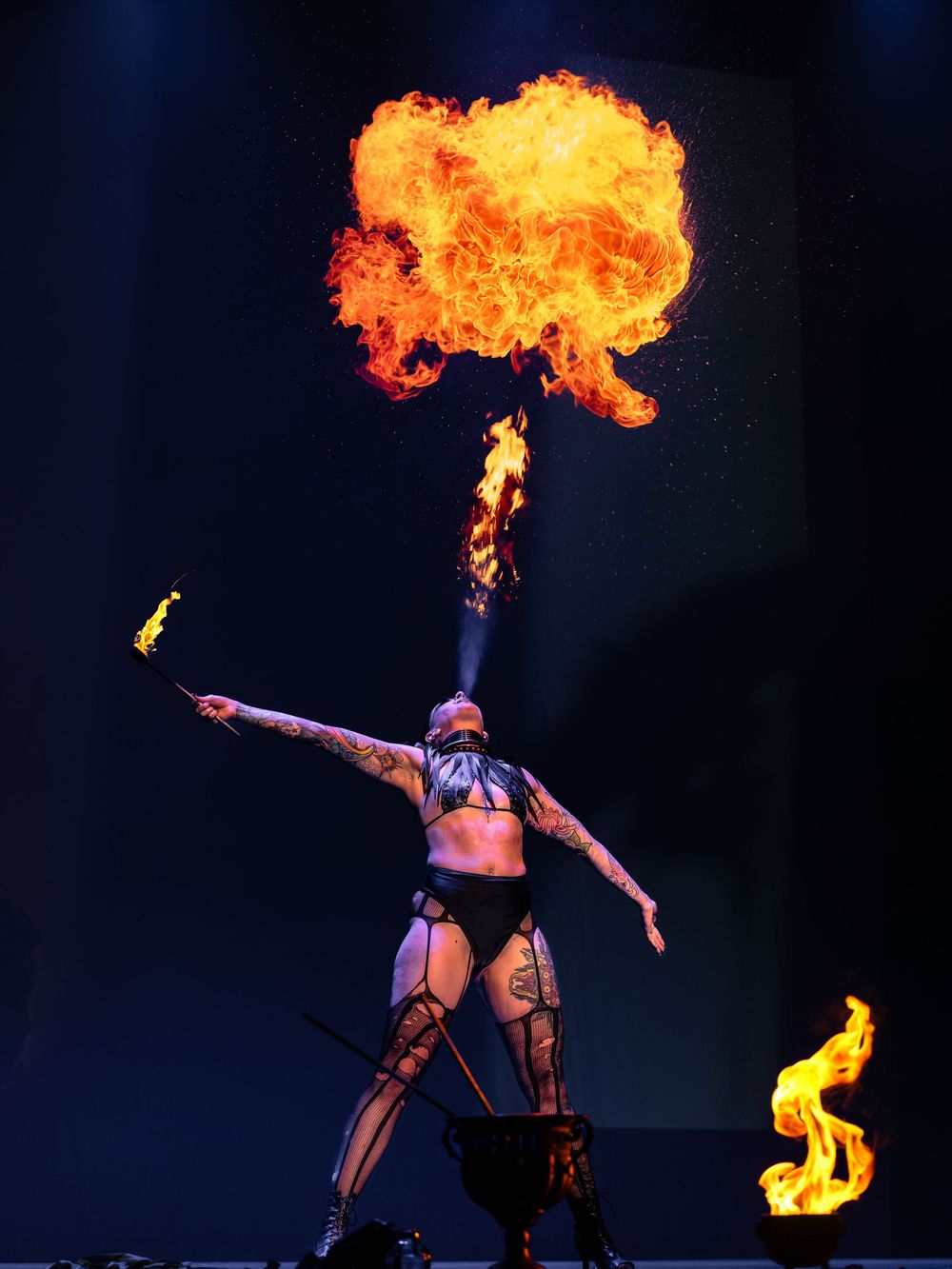 A performer in dark costume breathes fire creating a dramatic flame burst against a black background during a stage show.