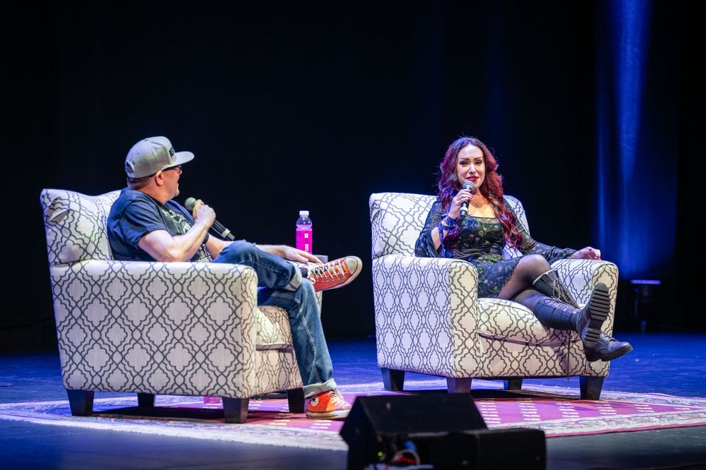 Two speakers engage in conversation while seated in patterned armchairs on a dimly lit stage with blue lighting.