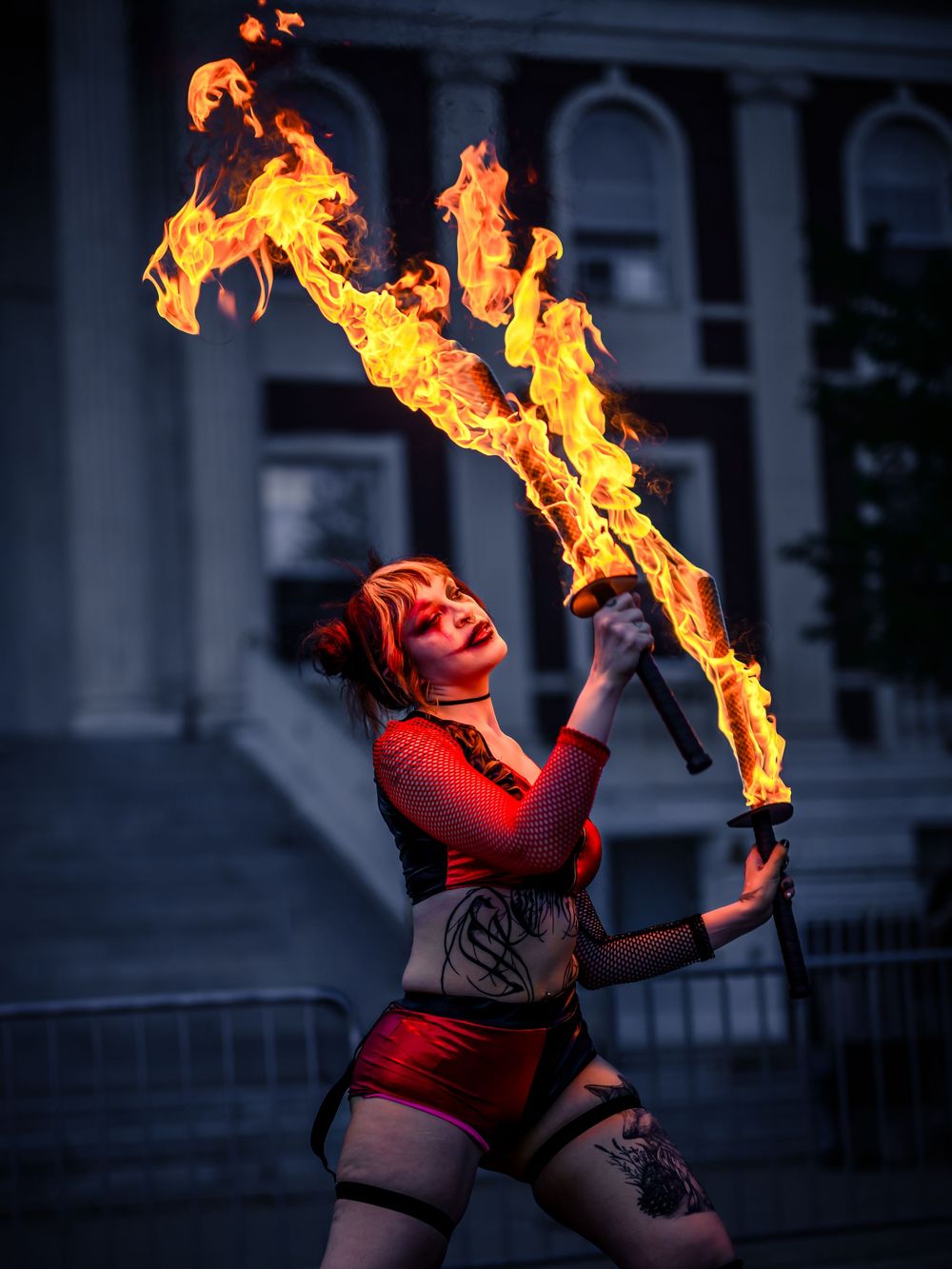 Female fire dancer performing with flaming batons outdoors at night in a red costume.