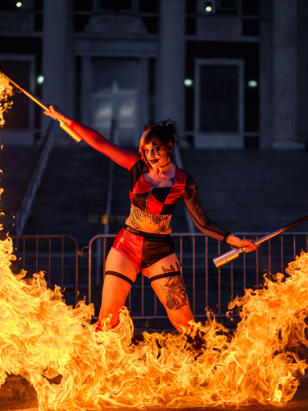Fire performer in red costume wielding flaming batons surrounded by large flames at night outdoor show.