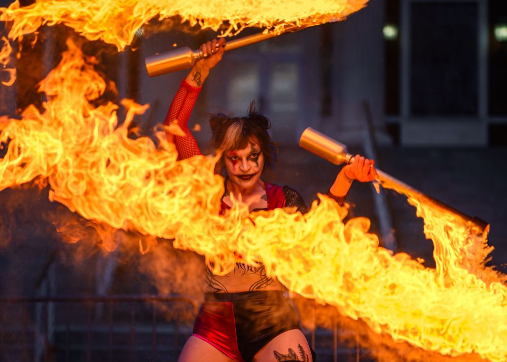 Fire performer in costume twirling flaming batons with dramatic orange flames blazing against dark background.
